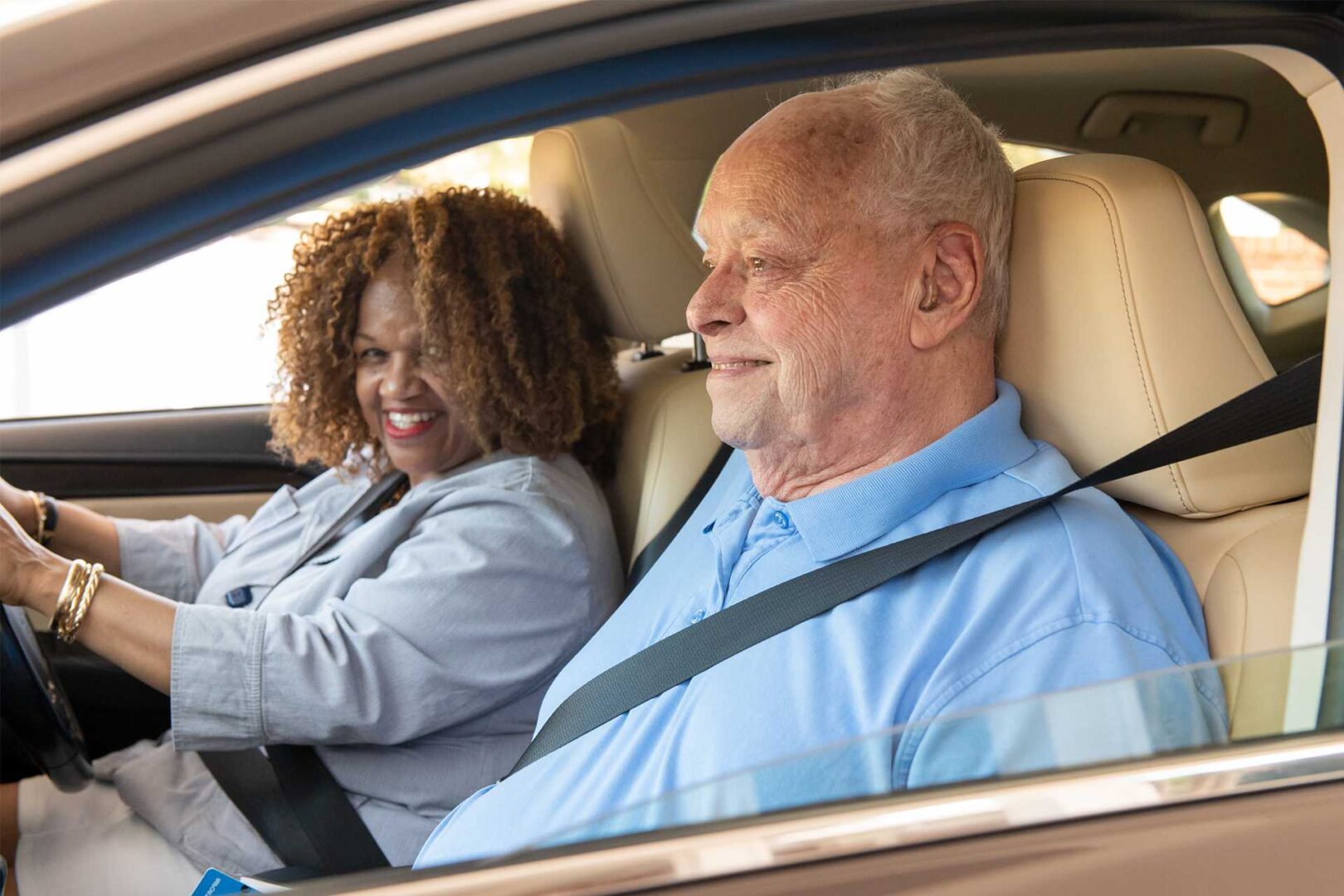 A young woman is the driver of a car with an elderly gentleman in the front passenger seat. Both are smiling because transportation for seniors is now solved!