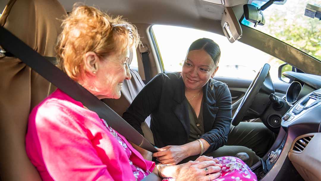 A young woman helps an elderly woman with her seat belt in the front seat of a car. They are enjoying the friendship they've developed through the NV Rides transportation partnerships. Both are happy because transportation for seniors is now solved!