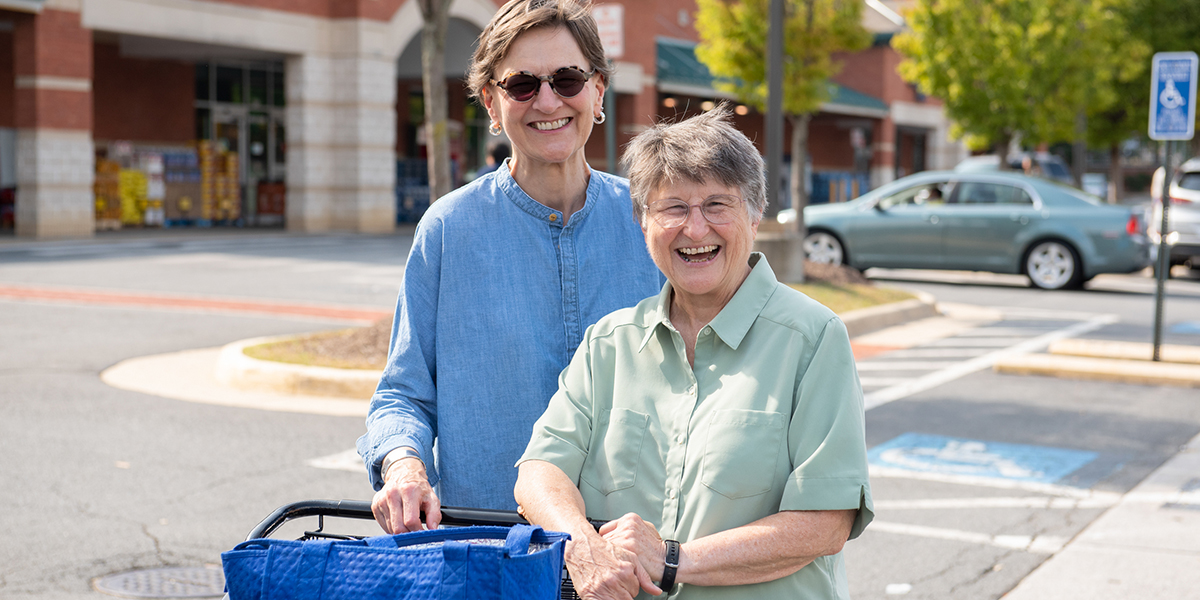 A woman helps another woman with putting groceries in the trunk of a car. They have become friends through a driving/riding partnership. Both are smiling because transportation for seniors is now solved!